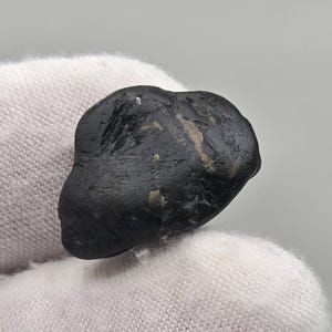 Op de afbeelding: Een donkere, onregelmatig gevormde steen met een ruwe textuur. De steen is voornamelijk zwart met enkele lichtere, bruinachtige insluitsels. Het object ligt op een witte, getextureerde ondergrond.