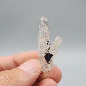 May include: A clear quartz crystal cluster with black inclusions. The crystal is double terminated and has a pointed shape.