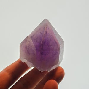 Könnte beinhalten: Ein facettierter Amethystkristall mit tiefviolettem Zentrum und helleren violetten Rändern. Der Kristall wird vor einem weißen Hintergrund in einer Hand gehalten. Die Facetten sind deutlich sichtbar.