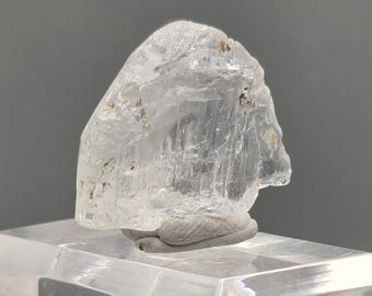 Op de afbeelding: Een helder, kristallijn mineraal specimen met een ruw, getextureerd oppervlak. Het mineraal is gemonteerd op een heldere acrylbasis. Het specimen heeft een lichte, doorschijnende uitstraling met enkele bruine insluitsels.