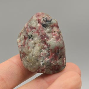 May include: A natural, uncut gemstone with a rough, textured surface. The stone features a mix of colors, including light gray, deep red, and black. The overall shape is irregular and organic, suggesting it is a raw, unpolished specimen. The stone is held in a hand.