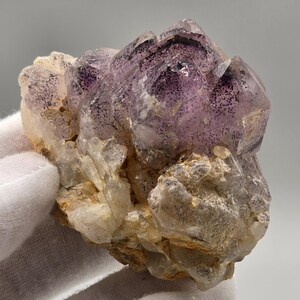 May include: A raw mineral specimen featuring translucent purple amethyst crystals clustered atop a base of jagged, off-white quartz. The purple crystal points are speckled with dark inclusions, creating a distinct contrast against the lighter, rough-textured mineral foundation.