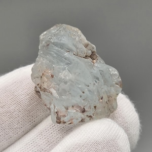 Peut inclure: Une pierre d'aigue-marine brute, de couleur bleu clair, avec une texture rugueuse et cristalline. Le minéral a un aspect translucide avec des inclusions brunes et beiges. La pierre est tenue dans une main gantée de blanc.