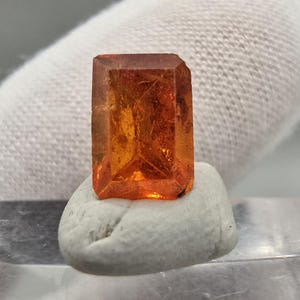 Peut inclure: Une gemme orange rectangulaire et facettée, possiblement un cristal, est présentée sur une petite base arrondie gris clair. La gemme a un aspect translucide, avec des structures internes visibles et une surface polie. L'éclairage met en valeur sa couleur et sa forme.