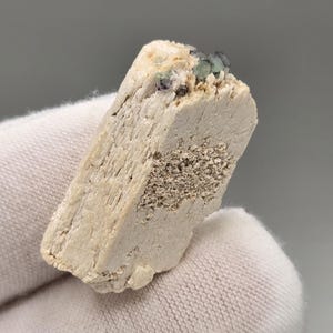以下が含まれることがあります： ざらざらとした質感の、明るいベージュ色の鉱物標本。上部には緑と紫の小さな結晶の塊が見える。手袋をはめた手で鉱物標本が持たれている。