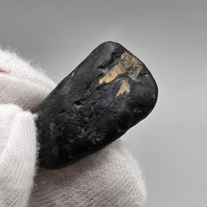 Op de afbeelding: Een kleine, onregelmatig gevormde zwarte steen met een ruw, getextureerd oppervlak. De steen heeft een opvallende, langwerpige goudbruine insluiting of ader in het bovenste gedeelte, die contrasteert met de donkere, matte basiskleur.