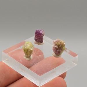 Op de afbeelding: Drie kleurrijke mineraalmonsters worden getoond op een heldere, vierkante acrylbasis. De mineralen omvatten een geelgroen, een dieppaars en een roze-groen kristal. Elk rust op een kleine, witte sokkel.