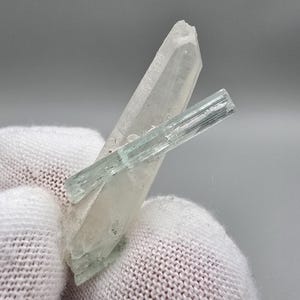 Peut inclure: Un cristal de quartz clair avec une petite formation cristalline bleu clair attachée. Le quartz est d'une couleur pâle et translucide, et le petit cristal a une forme rectangulaire. Les cristaux sont posés sur un fond neutre.
