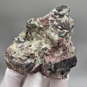 Könnte beinhalten: Ein raues, natürliches Mineralexemplar mit einer Mischung aus Farben. Der Stein weist Bereiche in Schwarz, Weiß, Rosa und Grün auf, wodurch eine strukturierte Oberfläche entsteht. Das Exemplar wird in einer behandschuhten Hand gehalten.