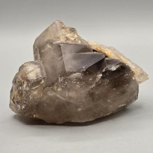 Peut inclure: Un amas de cristaux de quartz fumé. Le cristal est de couleur gris-brunâtre translucide avec une surface rugueuse et texturée. L'amas présente une structure géométrique complexe avec plusieurs faces cristallines bien définies.