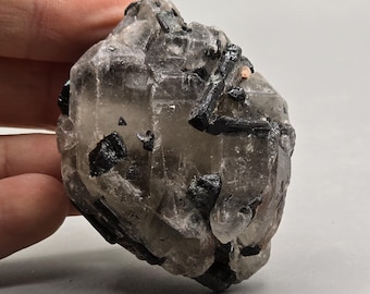 May include: A raw, natural crystal formation with a smoky quartz appearance. The crystal features embedded black tourmaline needles and a light brown hue. The rough texture and unique inclusions give it a striking, organic aesthetic.