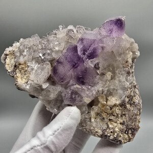 Pode incluir: Um aglomerado de cristais brutos de ametista e quartzo transparente com pontas roxas profundas e seções brancas translúcidas. O espécime mineral exibe inclusões naturais e manchas escuras por toda a estrutura cristalina, fixado em uma base rochosa rugosa.