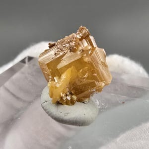 May include: A golden-yellow wulfenite crystal specimen featuring a distinct cubic structure with sharp, translucent edges and a cluster of smaller, lighter yellow crystals attached to the base, displaying a natural, multifaceted mineral formation.
