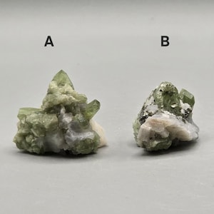May include: Two raw, uncut green peridot crystal clusters, labeled A and B, sit on a gray surface. The crystals have a light green hue with some white and gray matrix. The crystals are rough and natural.