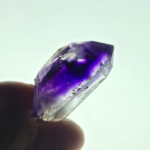 Könnte beinhalten: Ein roher Amethystkristall mit tiefvioletter Spitze, der allmählich in eine durchscheinende, hellviolette und klare Basis übergeht. Der Kristall wird in einer Hand gehalten und zeigt seine natürliche, unpolierte Form. Die Facetten des Kristalls fangen das Licht ein.