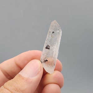 May include: A clear quartz crystal with black inclusions. The crystal is pointed at the top and has a smooth, polished surface.