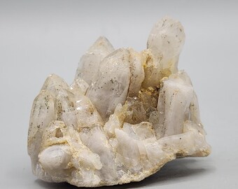 Puede incluir: Un grupo de cristales de cuarzo transparente con una textura natural y rugosa. Los cristales tienen un color blanco roto pálido con sutiles inclusiones y variaciones. La formación de cuarzo tiene múltiples puntas y una forma orgánica y robusta.