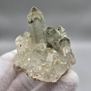 May include: A cluster of clear quartz crystals featuring a prominent central point with internal phantom inclusions and dark mineral deposits. The specimen displays multiple smaller, sharp-edged crystalline formations radiating from the base.