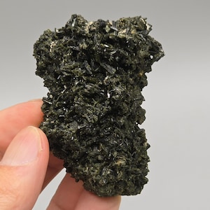 Peut inclure: Spécimen minéral vert foncé avec une surface rugueuse et texturée. Le minéral est composé de nombreux petits cristaux en forme d'aiguilles, créant un aspect épineux. La couleur est un vert forêt profond avec quelques zones plus claires.