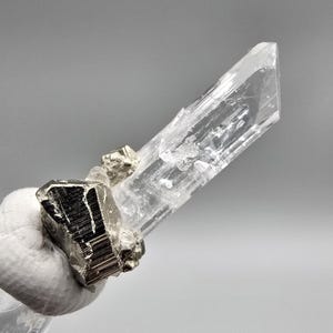 May include: A clear, elongated crystal with a faceted tip, alongside several metallic, dark-coloured mineral formations. The crystal is partially translucent, revealing internal structures. The minerals are set against a neutral grey background.
