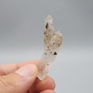 May include: A clear quartz crystal with black inclusions. The crystal is pointed at the top and has a rough, textured surface.