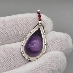 Peut inclure: Un pendentif en forme de goutte d'eau avec une pierre précieuse violet foncé polie, sertie dans un cadre argenté. Le haut du pendentif présente une petite barre ornée de plusieurs petites pierres précieuses rouges. Le pendentif est tenu sur un fond blanc.