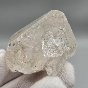 Peut inclure: Un amas de cristaux de quartz clair avec plusieurs pointes. Le cristal est tenu dans une main gantée, mettant en valeur son aspect naturel et translucide. Le quartz a une surface rugueuse et texturée.