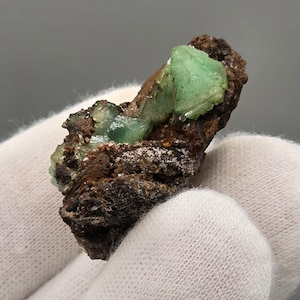 Puede incluir: Primer plano de un espécimen mineral con cristales verdes translúcidos vibrantes sobre una matriz marrón rugosa. Los cristales tienen una forma angular distinta, que contrasta con la textura terrosa de la base. El espécimen se sostiene en una mano enguantada de blanco.