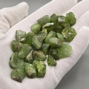 May include: A collection of raw peridot gemstones in various sizes and shapes. The stones are a vibrant, translucent green with brown and tan patches. The peridot is held in white fabric.