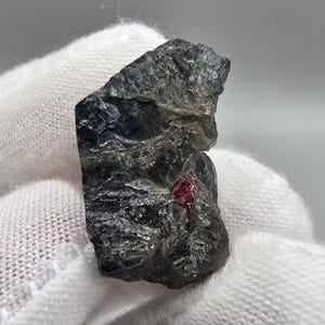 Peut inclure: Spécimen minéral brut de couleur gris foncé avec un petit cristal rouge vif incrusté. La pierre a une surface texturée et est tenue par une main gantée de blanc. L'aspect général suggère une pierre précieuse brute et naturelle.