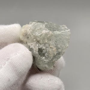 May include: A pale blue-green mineral specimen with a rough, crystalline structure. The stone has a translucent quality, with areas of white and light brown inclusions. The mineral is held by a gloved hand.