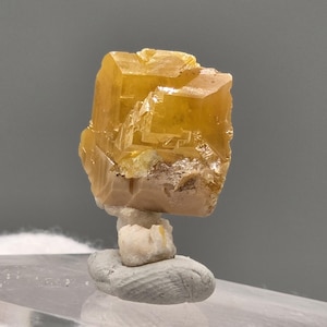 May include: A translucent golden-yellow wulfenite crystal specimen featuring sharp, blocky, tabular geometry with a vitreous lustre. The mineral displays complex stepped growth patterns and sits atop a small, light-coloured, textured matrix base.