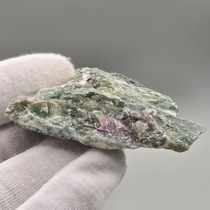 Peut inclure: Un spécimen minéral brut présentant une surface rugueuse et texturée avec un mélange de tons vert forêt profond et olive. Des taches distinctes de magenta et de rose vifs sont incrustées dans la structure cristalline, créant un contraste.