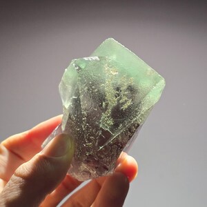 Puede incluir: Un cristal de fluorita verde con una textura clara y vítrea. El cristal tiene una superficie natural, rugosa y es parcialmente translúcido.