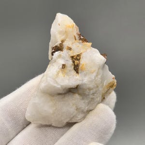 Peut inclure: Spécimen de cristal de quartz brut et naturel. Le quartz est principalement blanc avec des inclusions minérales brunes et beiges. Le cristal est tenu dans une main gantée, mettant en valeur sa surface rugueuse et texturée et ses caractéristiques géologiques uniques.