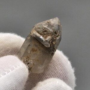 Peut inclure: Une pointe de cristal de quartz translucide aux tons fumés présentant une terminaison naturellement facettée. La surface affiche des inclusions minérales terreuses et brunes ainsi qu'une petite croissance cristalline secondaire.