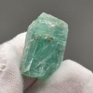 Peut inclure: Un cristal brut, non taillé, de couleur aigue-marine. La pierre précieuse présente une surface rugueuse et texturée avec des structures cristallines visibles et de subtiles variations de couleur. Le cristal est tenu sur un fond de tissu blanc.