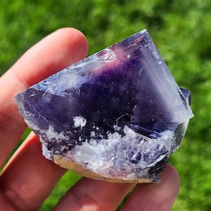 Puede incluir: Un gran cristal de fluorita púrpura con un acabado claro y brillante. El cristal tiene una forma geométrica afilada con muchas facetas. La fluorita es una gema natural.