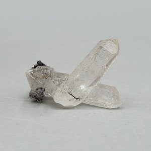 May include: A clear quartz crystal with a black mineral inclusion. The crystal is double-terminated and has a pointed shape.