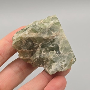 Peut inclure: Spécimen minéral brut de couleur vert clair, avec une structure cristalline. La pierre a une forme naturelle et irrégulière et un aspect légèrement translucide. La couleur varie du vert pâle au vert moyen, avec des zones plus opaques.