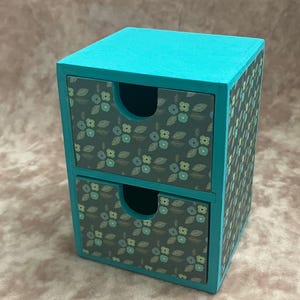 May include: A small wooden storage unit with two drawers, featuring a bright turquoise frame and drawer fronts decorated with a dark grey floral pattern of light blue and green flowers and leaves, each drawer has a circular cutout for opening.