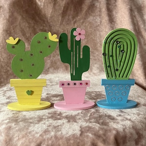 May include: Three wooden cactus ornaments. The cactus on the left is green with yellow flowers, the middle cactus is green with a pink flower, and the right cactus is green with small black accents. Each cactus sits in a colourful pot.