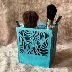 May include: A turquoise, square makeup brush holder with a cut-out leaf and butterfly design. The holder contains several makeup brushes with black and brown bristles. The holder is made of wood and has a matte finish.