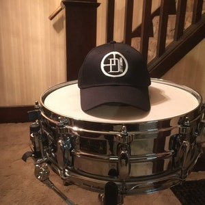 Op de afbeelding: Een zwarte baseballpet met een wit logo met een cirkel met de letter "D" erin. De pet ligt op een zilveren snaredrum.