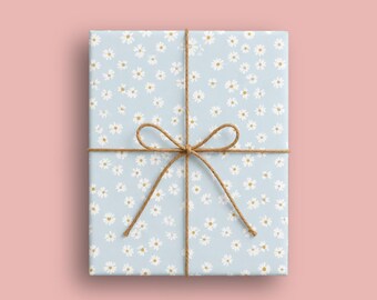 Op de afbeelding: Een rechthoekige geschenkdoos verpakt in lichtblauw papier met een herhalend patroon van kleine witte madeliefjes met gele hartjes. Het pakket is omwikkeld met een dun, natuurlijk bruin touw in een kruispatroon met een strik.