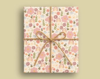 Op de afbeelding: Een rechthoekige geschenkdoos verpakt in crèmekleurig papier met een herhalend patroon van roze bloemen, oranje regenbogen, groene bladeren en kleine geel-zwarte bijen, vastgebonden met een dun bruin touwtje in een strik in het midden.