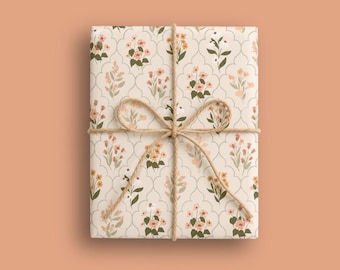 Op de afbeelding: Een rechthoekig cadeau verpakt in gebroken wit papier met een herhalend patroon van kleine roze en oranje bloemen met groene bladeren in een dun grijs raster. Het pakket is vastgebonden met een eenvoudige bruine touwstrik in het midden.