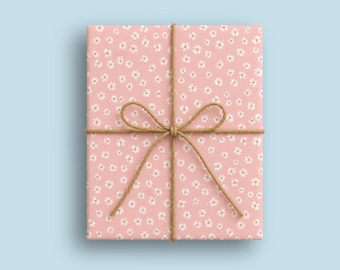 Op de afbeelding: Een rechthoekige geschenkdoos verpakt in lichtroze papier met een herhalend patroon van kleine witte bloemetjes die op madeliefjes lijken. Het pakket is vastgebonden met een dun, natuurlijk bruin jutekoord in een kruispatroon met een strik.