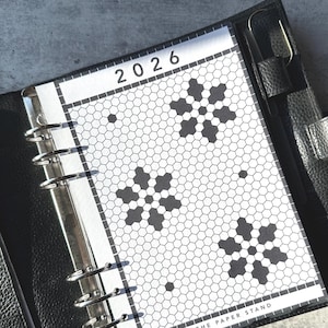 May include: Black leather planner with a 2026 calendar insert. The insert features a white background with a black honeycomb pattern and snowflake designs. The text "THE PAPER STAND" is visible at the bottom.
