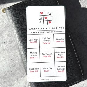May include: A Valentine's Day Tic-Tac-Toe game card with a black leather wallet. The card features a tic-tac-toe grid with date night ideas like "Movie Night" and "Stargazing". The text "Valentine Tic-Tac-Toe" is at the top.